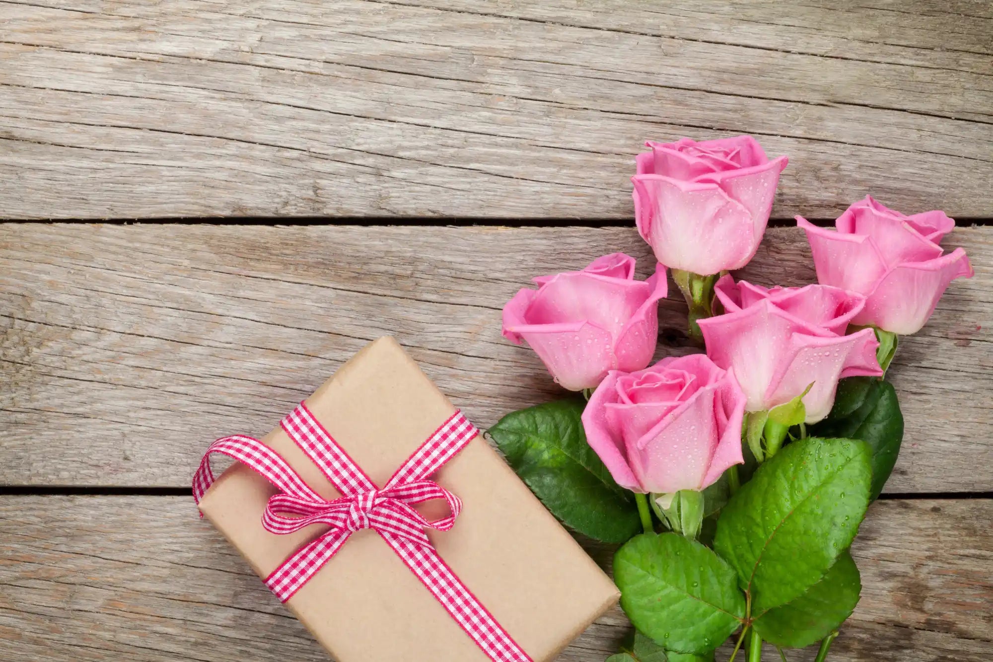 Bouquet of pink roses next to a gift wrapped in kraft paper with a gingham ribbon.