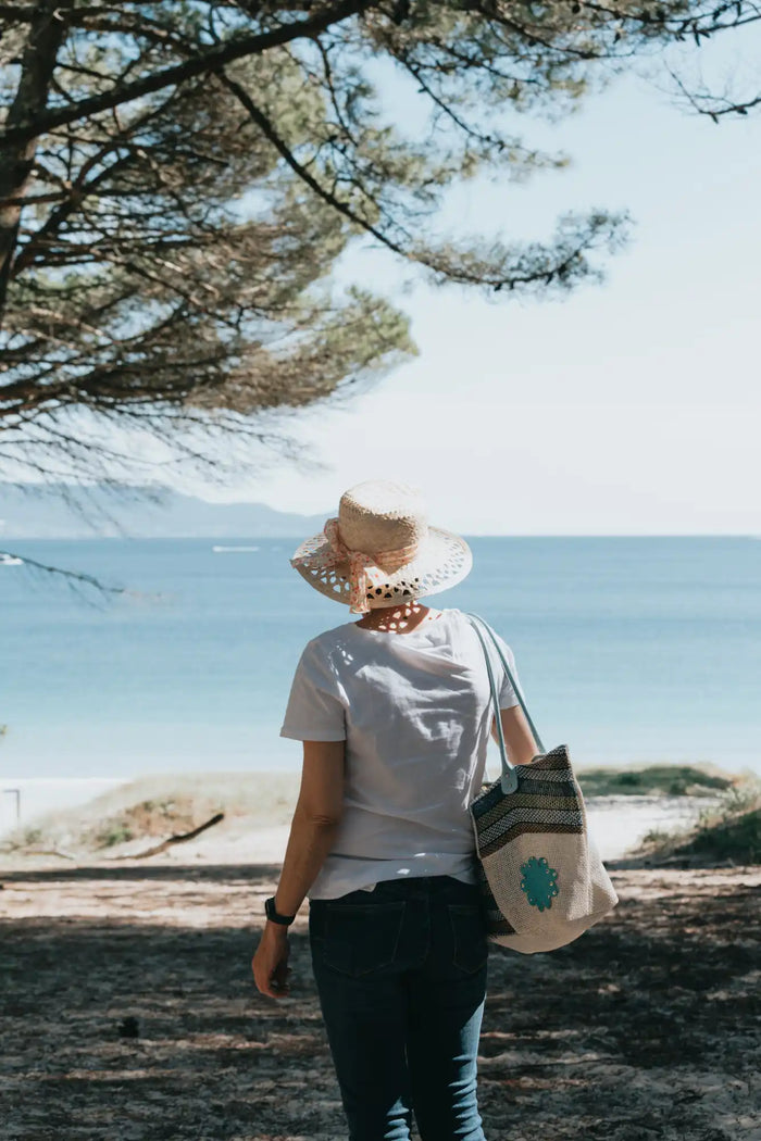 Person wearing a sun hat and carrying a tote bag, standing near a beach.